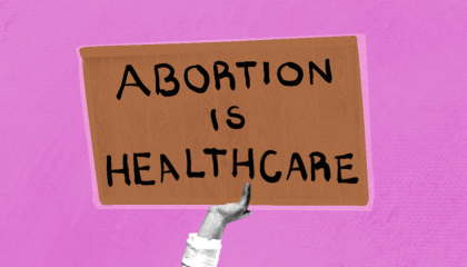 On a pink background, a hand holds up a brown sign reading, "Abortion is healthcare."
